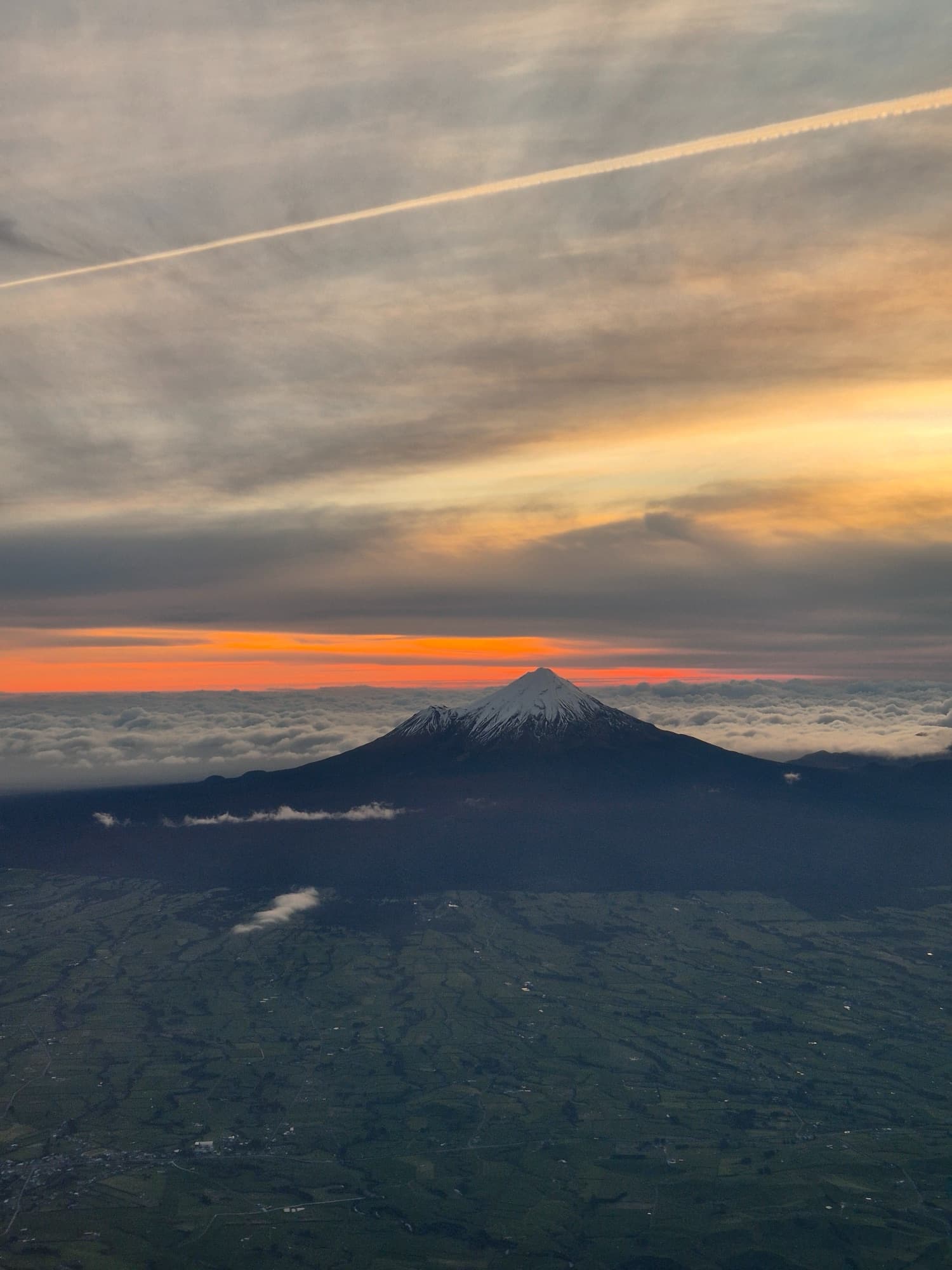 Aerial view of Maunga Taranaki at sunset, its snow-capped summit rising above the clouds over the Taranaki plains